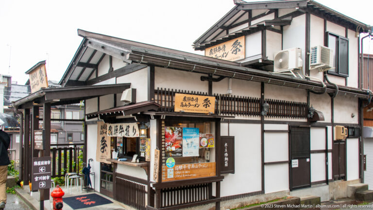 Black-and-white traditional corner building with wooden signs, food counter, balcony, menus, and colorful posters.
