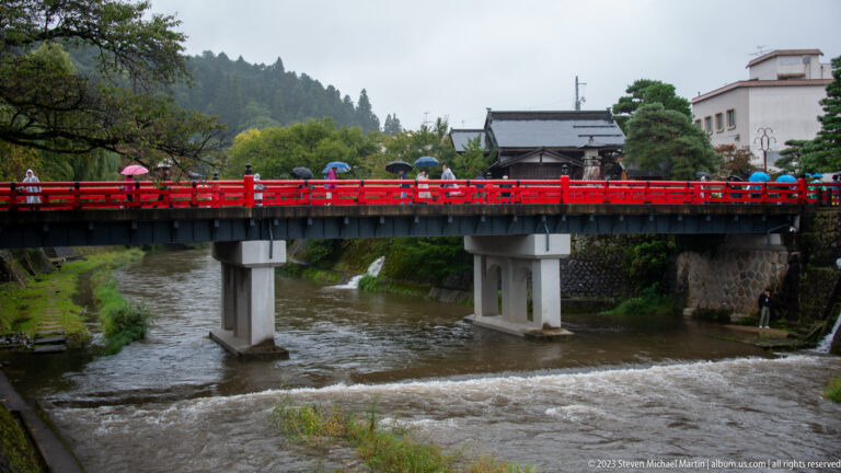 Bright red bridge crosses a swollen river as umbrella-carrying pedestrians pass between traditional buildings and wooded hills.