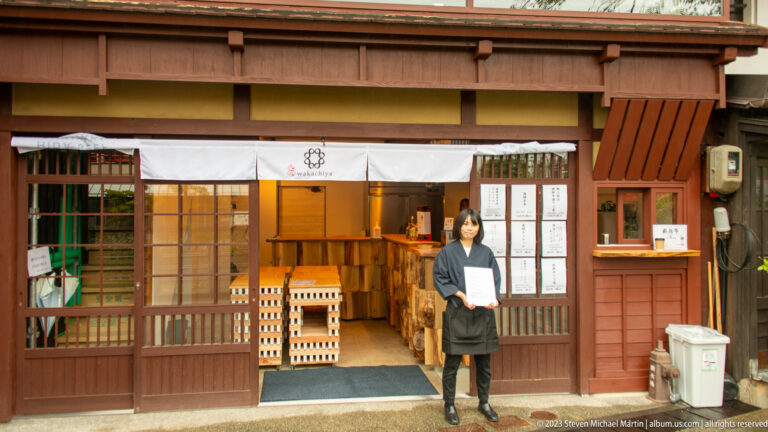 Woman standing at the entrance of an open wooden craft shop with shelves and pale timber goods inside.