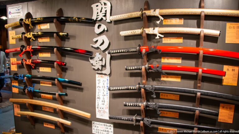 Rows of Japanese-style swords with black, red, blue, and wood-toned scabbards fill a shop display wall.