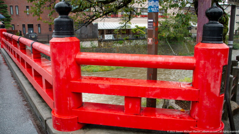Close view of vermilion bridge rails and posts with black finials beside a river and wet pavement.