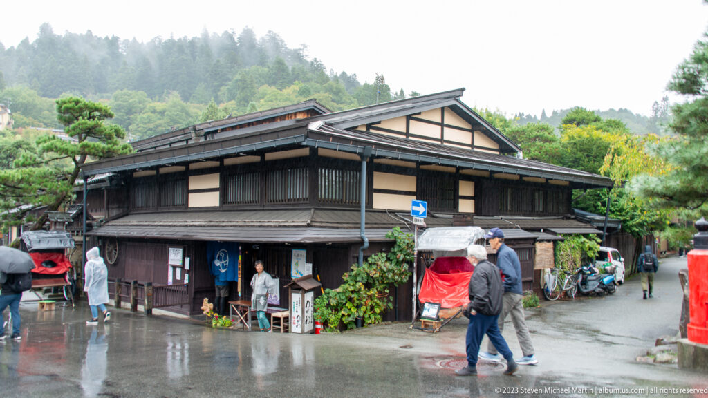Large dark wooden townhouse at a rainy street corner with pedestrians, umbrellas, and a red rickshaw.