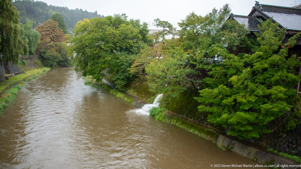 Brown river flows between green embankments, with a small outfall and traditional roofs partly visible through trees.