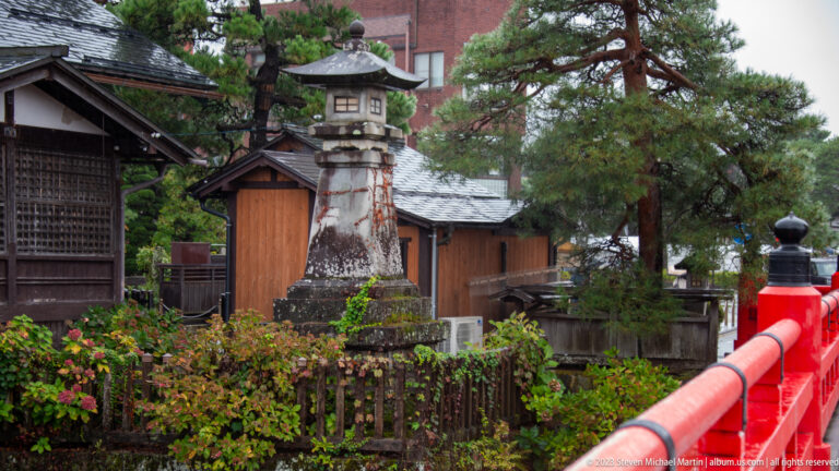 Weathered stone lantern stands beside red bridge railing among pine branches, shrubs, and autumn foliage.