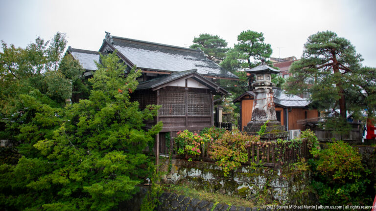 Traditional timber buildings and layered roofs stand behind a riverside garden with stone lantern, pines, shrubs, and fence.