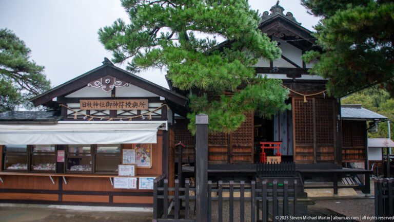 Gabled timber religious building with lattice doors, visitor counter, Japanese signboard, pine branches, and red structure beyond.