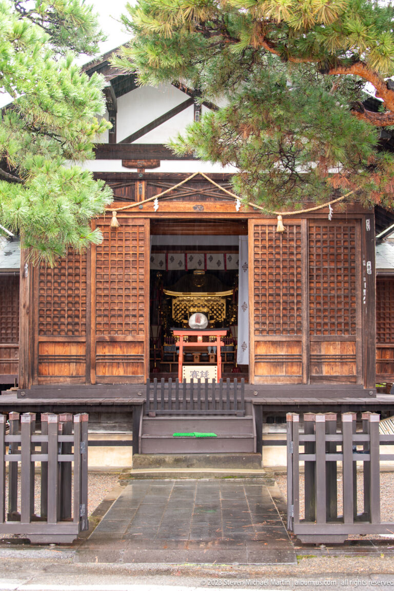 Open wooden Shinto worship hall with central offering area, sacred rope, paper streamers, steps, and symmetrical railings.