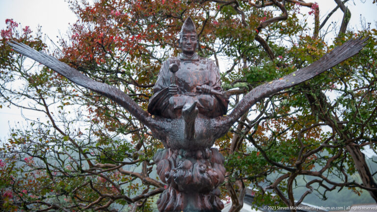 Bronze Japanese figure in formal attire rides a large winged bird against autumn leaves and pale sky.
