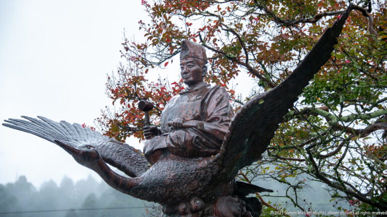 Side view of bronze courtly figure riding a long-winged bird, with autumn branches and misty hills behind.