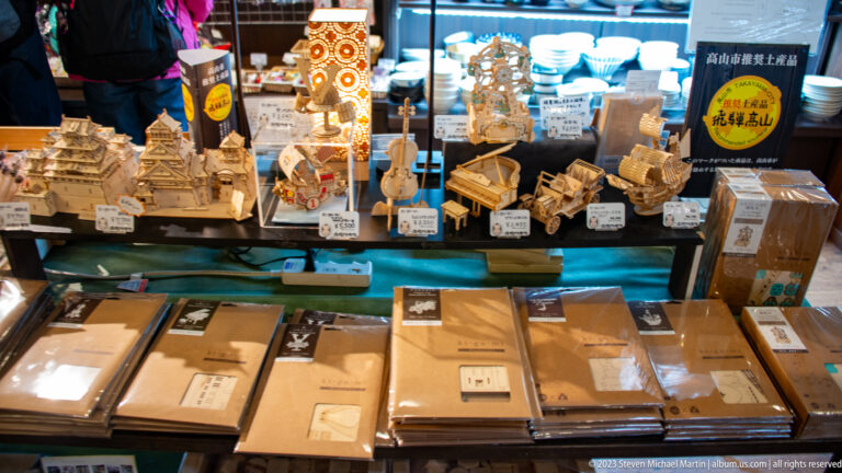 Miniature wooden buildings, carts, decorative objects, and craft kits are arranged in a brightly lit shop display.