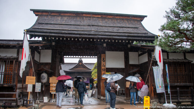 Large timber gate with broad roof, Japanese flags, banners, and umbrella-carrying visitors entering a historic compound.