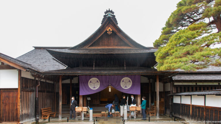 Large timber hall with steep gable, purple curtain bearing paired crests, white walls, and visitors at the entrance.