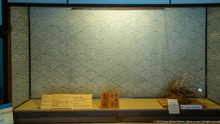 Museum display case with small labeled objects before a repeating blue-and-gold Japanese wave pattern.