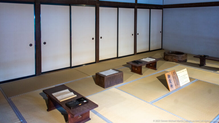 Tatami room with low writing desks, document boxes, small stands, and plain sliding wall panels.