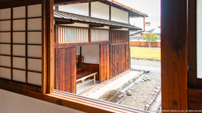 Open wooden window frames a gravel courtyard, low timber service building, bench, and stone-edged path.