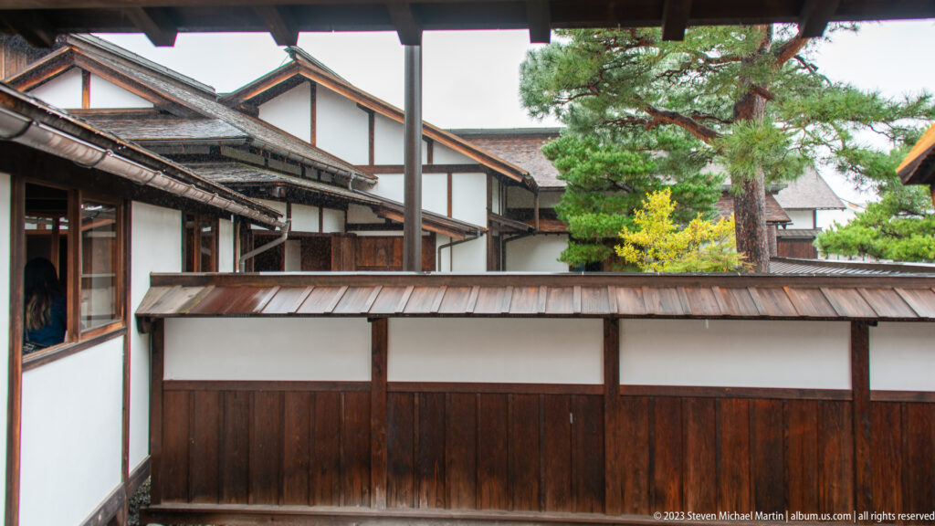 White-plaster and dark-timber courtyard walls with low metal roofs, pine tree, yellow shrub, and surrounding corridors.