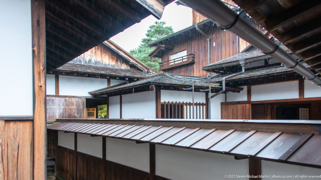 Layered historic roofscape with overlapping eaves, gutters, white-plaster walls, timber buildings, and enclosed corridors.