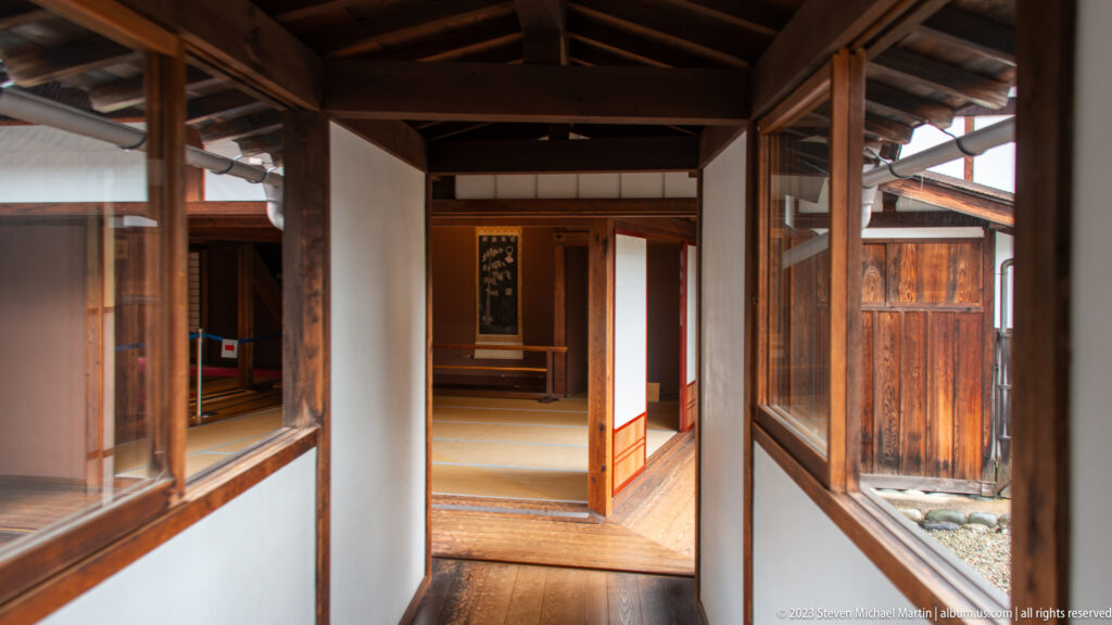 Narrow timber corridor with polished floorboards, glazed openings, exposed beams, and a tatami room with hanging scroll beyond.