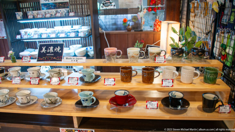 Ceramic cups, bowls, and small vessels in varied glazes line a wooden shop counter with individual price tags.