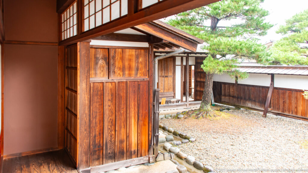 Mature pine stands in a gravel courtyard enclosed by timber walls, sliding openings, stones, and low garden edging.