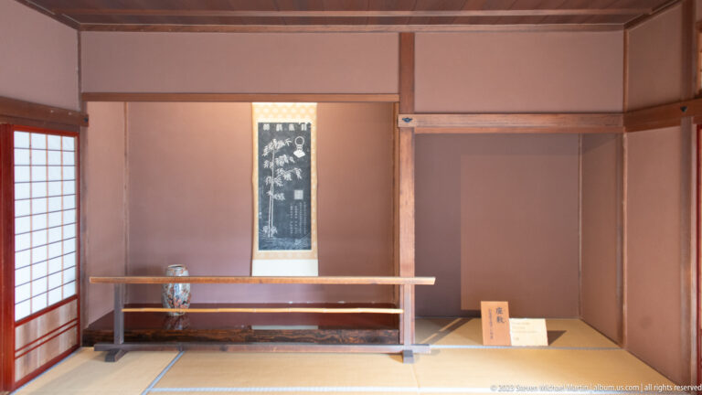 Sparse tatami room with hanging calligraphy scroll, low wooden rail, small ceramic vessel, and pale plaster walls.