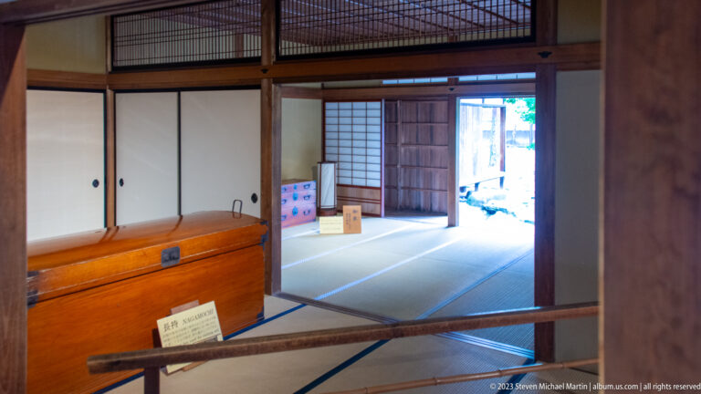 Tatami room with sliding panels, exposed wood framing, built-in cabinetry, and an open doorway to another space.