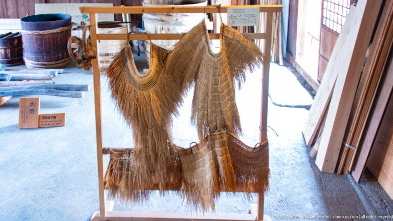 Traditional Japanese straw rain cape hangs on a wooden frame, with broad shoulders and long layered fringe.