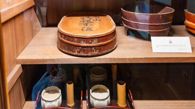 Stacked round lacquered food containers and a large bowl sit on a wooden shelf above smaller vessels.