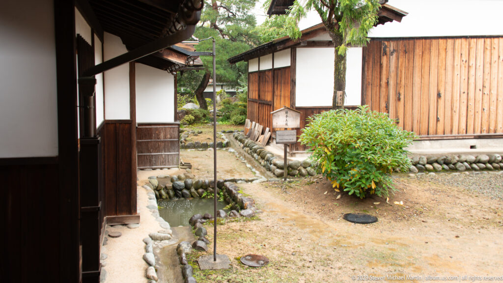 Earthen service yard with timber outbuildings, white-plaster walls, stone edging, drainage channel, shrub, and narrow paths.
