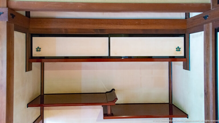 Tokonoma-style alcove with staggered wooden shelves, recessed platform, pale sliding panels, and small floral crests.