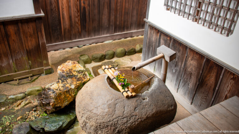 Large rounded stone water basin with bamboo spout, small flower arrangement, mossy stones, and timber wall.