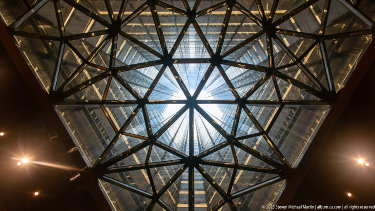 Upward view of a faceted glass ceiling with dark triangular steel framing and bright perimeter spotlights.