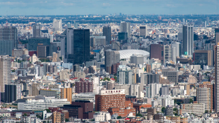 Telephoto city view with the white curved roof of Tokyo Dome among dense buildings and office towers.