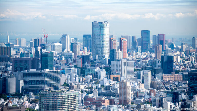Telephoto Tokyo skyline with a tall glass tower, office buildings, cranes, and dense urban development.