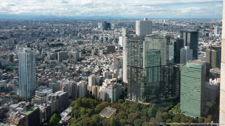 Glass and concrete towers beside wooded open space, with dense Tokyo neighborhoods beyond.