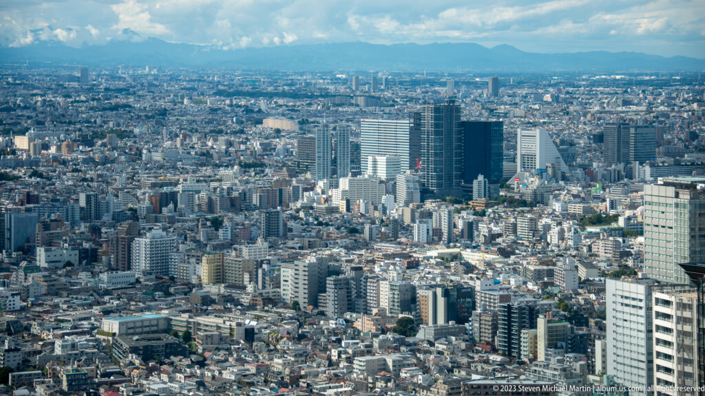 Western Tokyo cityscape with modern towers and a distant mountain ridge beneath clouds.