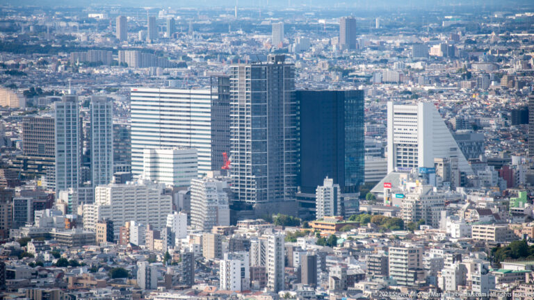 Telephoto view of white, blue-glass, and angular office towers surrounded by lower Tokyo buildings.