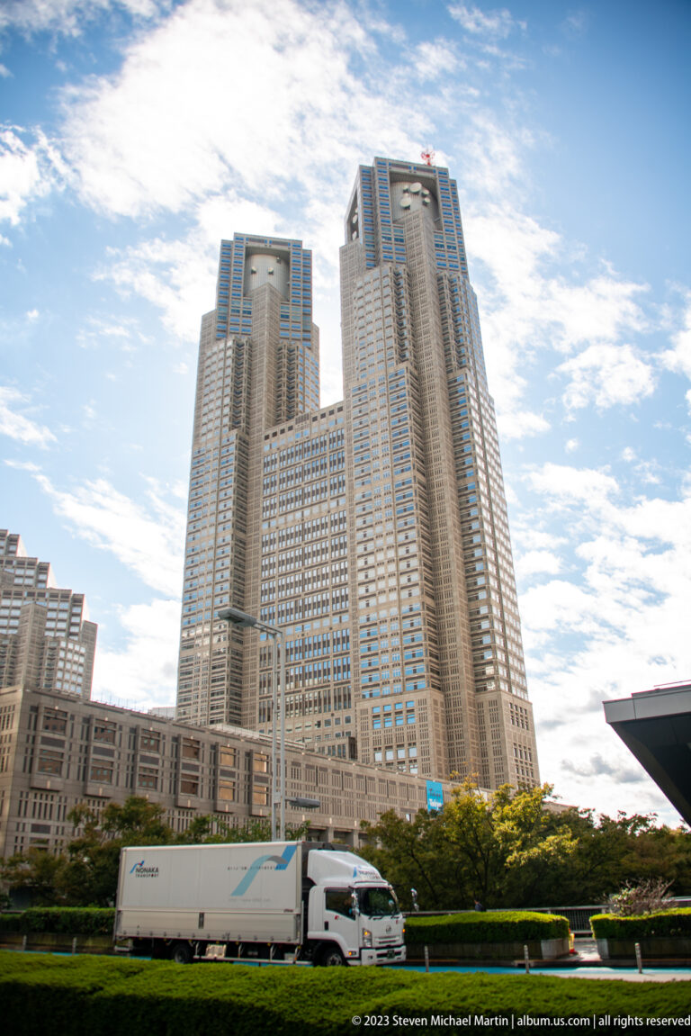 Low-angle street view of the twin-towered Tokyo Metropolitan Government Building above trees and a passing truck.