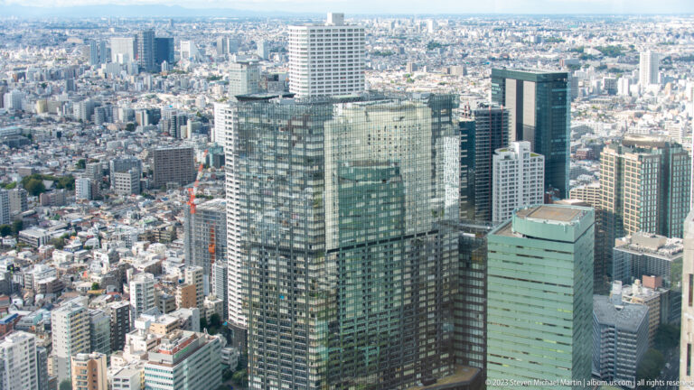 Close elevated view of a broad glass tower with a rooftop volume and a green tower beside it.