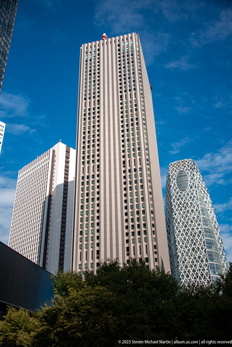 Low-angle Shinjuku skyline with rectangular towers and the white-latticed Mode Gakuen Cocoon Tower at right.