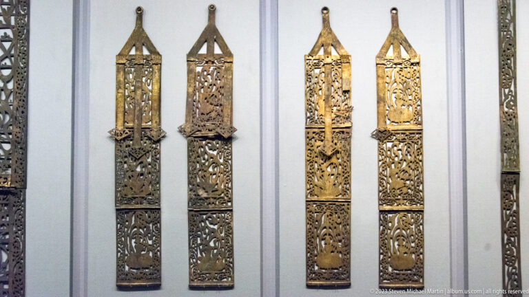 Four long openwork gilt-bronze pendants with triangular loops displayed beside narrow decorative strips.