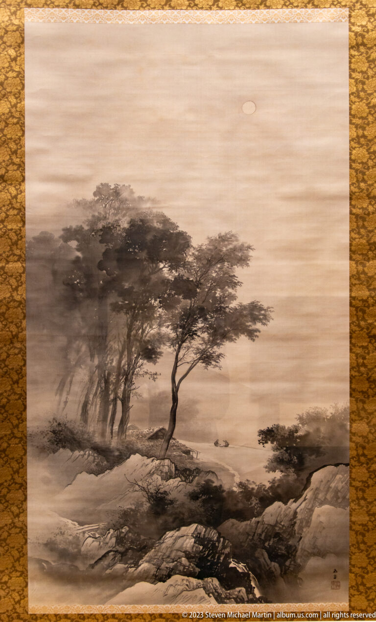 Monochrome ink landscape with windswept trees, rocky foreground, water, and pale sky.