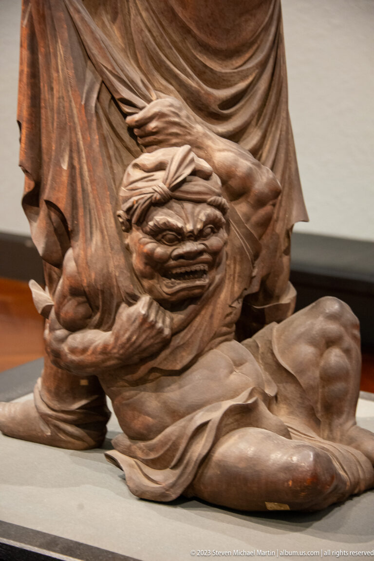 Close-up of a carved wooden guardian’s tense hand resting against drapery and a bent knee.