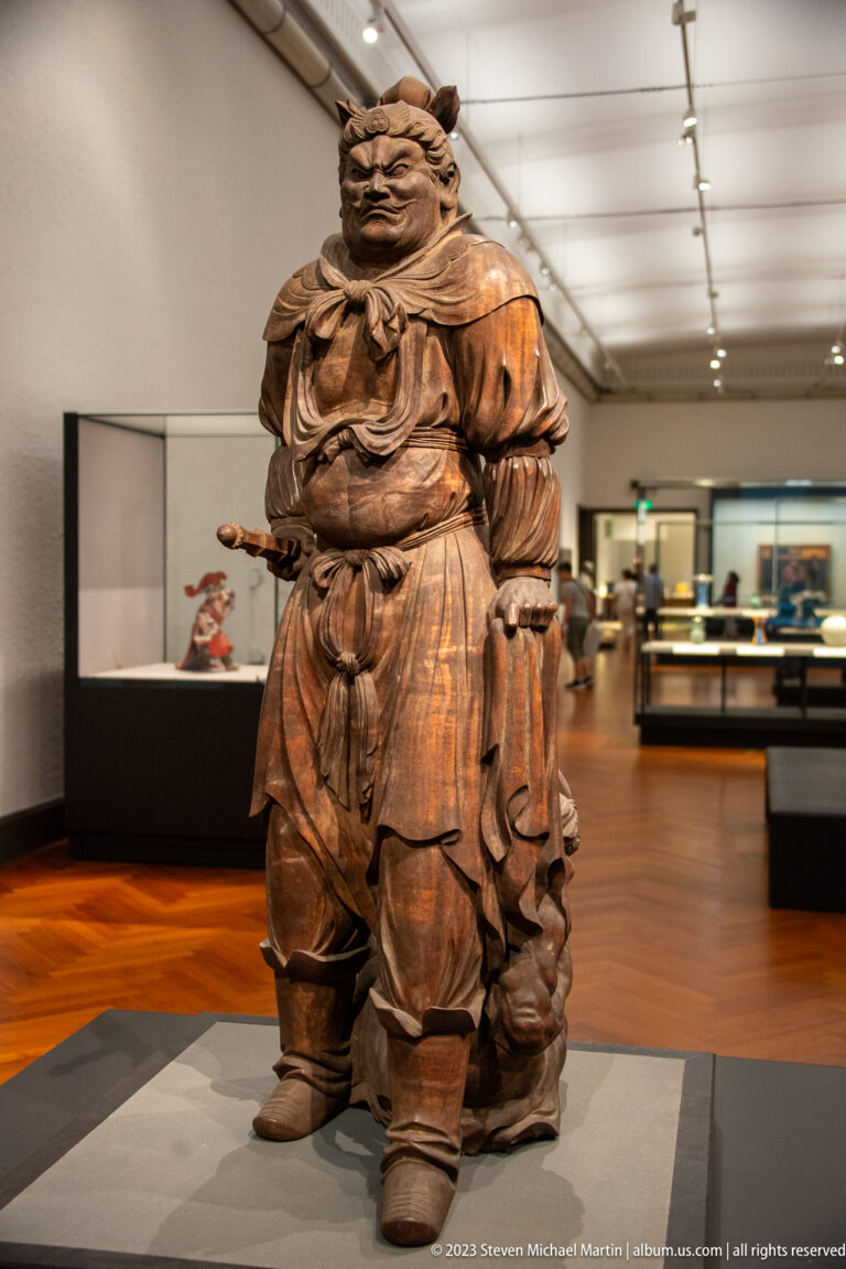 Large standing wooden Buddhist guardian with stern face, layered armor, and deeply carved robes.