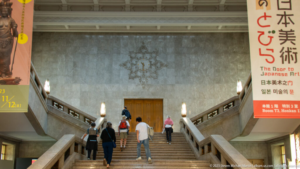 Visitors ascending the Honkan’s broad central staircase beneath tall museum exhibition banners.