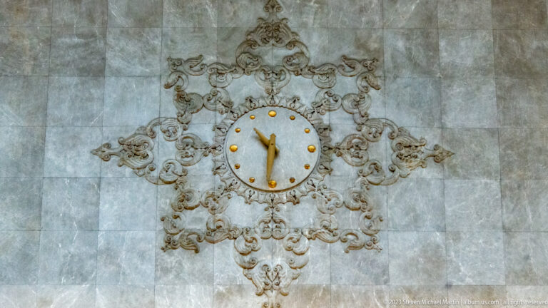 Round wall clock with gold hands centered in an ornate symmetrical relief on pale stone.