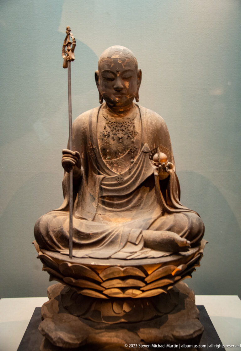 Seated wooden Buddhist monk holding a tall staff and small ritual object on a lotus base.