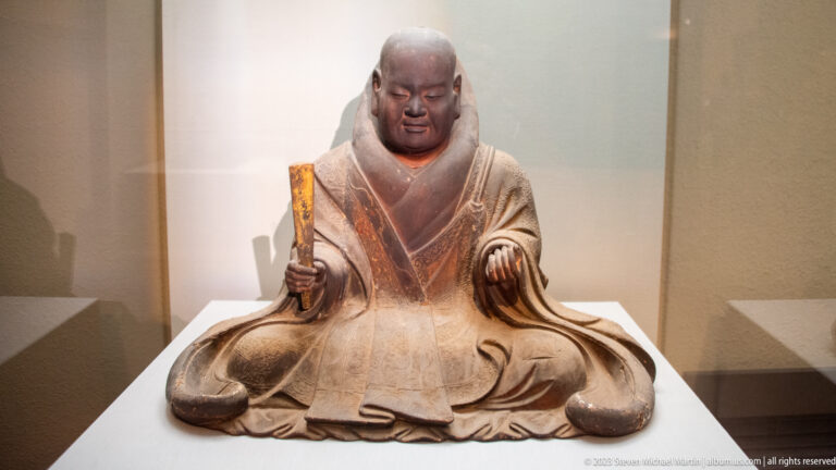 Polychrome wooden seated monk holding a narrow rectangular tablet and extending one hand.