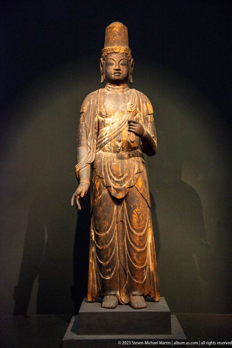 Standing polychrome wooden Buddhist figure with tall cap, layered robes, and one hand across the chest.