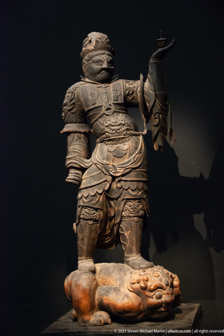 Armored wooden Buddhist guardian standing on a prostrate demon-like figure.
